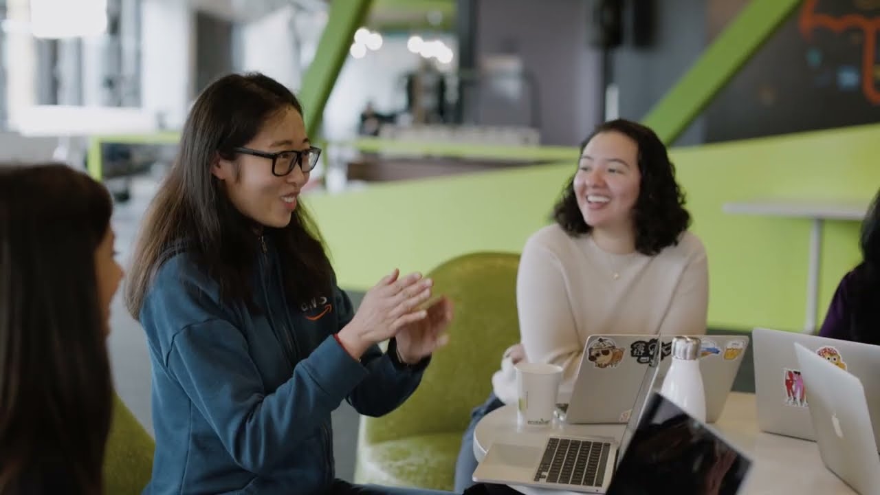 A group of people having a discussion around a table in a modern, casual office environment. Laptops, drinks, and casual attire suggest a collaborative and friendly workplace atmosphere.