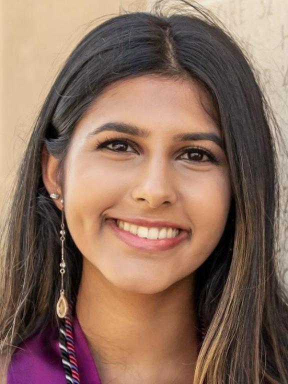 Portrait of a smiling woman with long dark hair, wearing a long earring, in front of a neutral background.