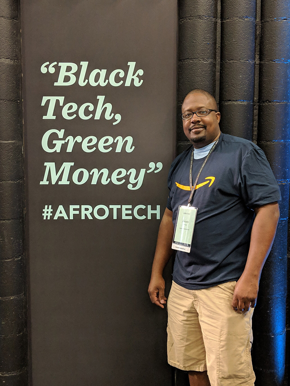 A person wearing an Amazon shirt stands next to a sign that reads 'Black Tech, Green Money' with the hashtag #AfroTech, likely at a tech or diversity-focused event.