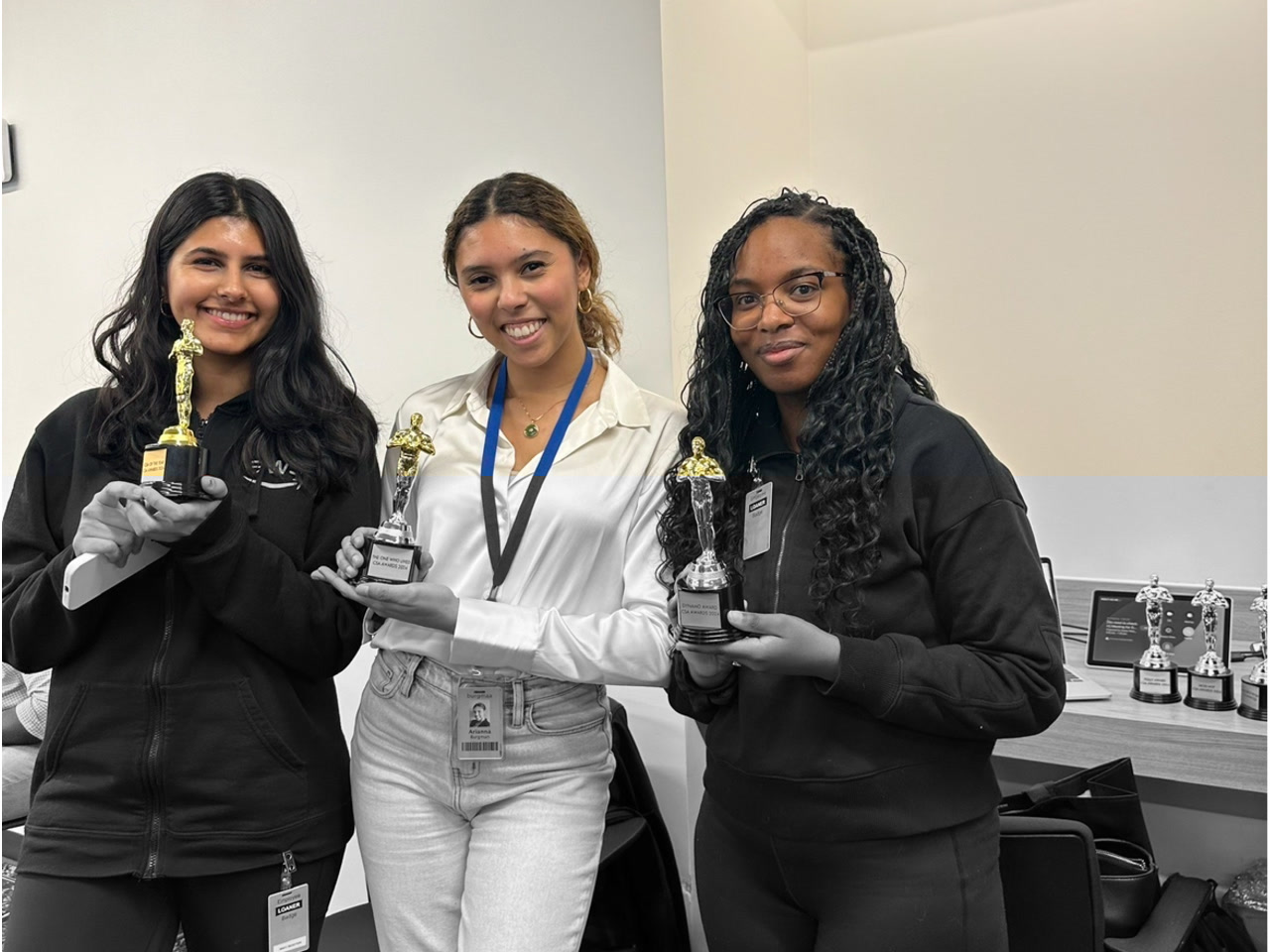 Three women standing and holding award trophies in an office setting, smiling for the camera. Additional trophies and office materials are visible in the background.