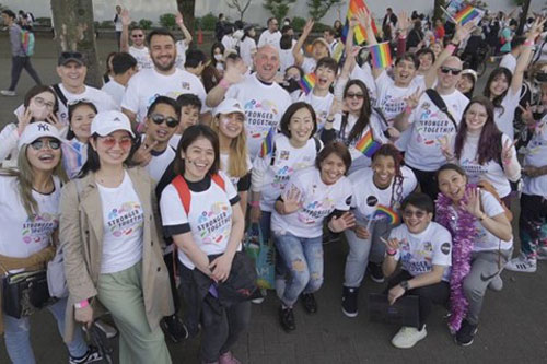 A diverse group of people wearing matching shirts pose together at a Pride event, celebrating with rainbow flags and smiles.