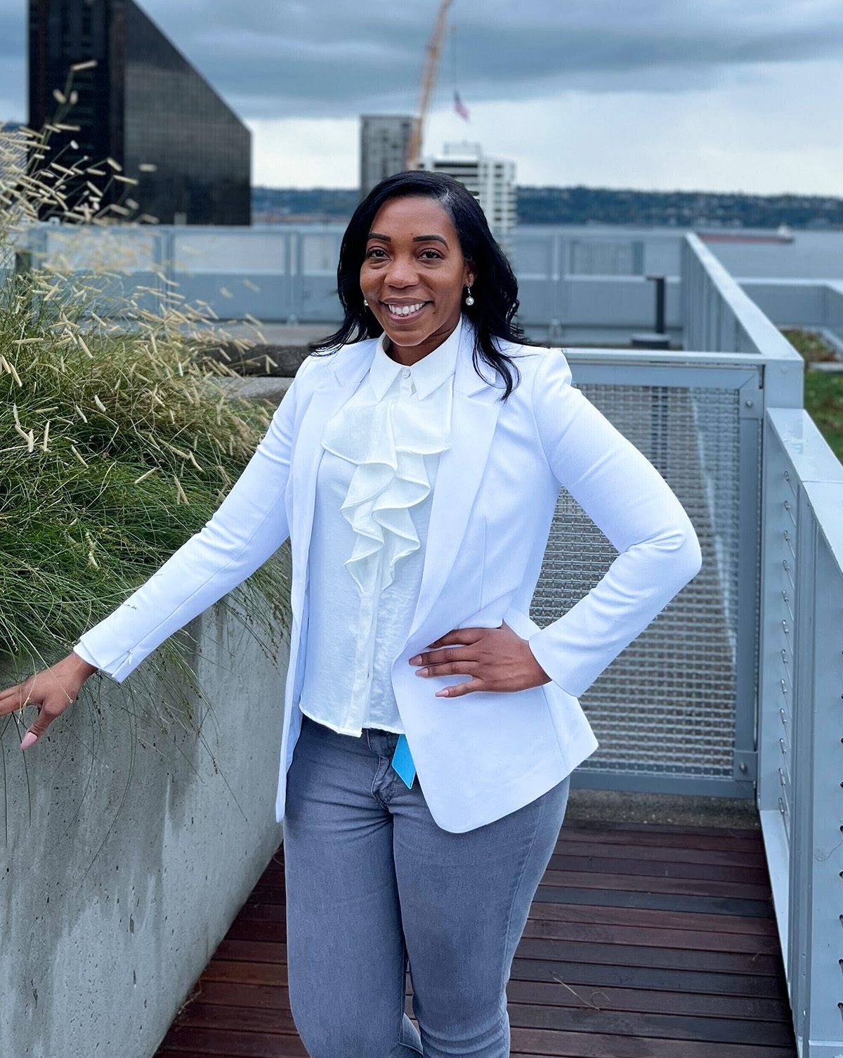 A woman stands outdoors on a rooftop or terrace with an urban background, wearing a white blazer and a ruffled blouse, smiling at the camera. The setting includes modern buildings and waterfront scenery.