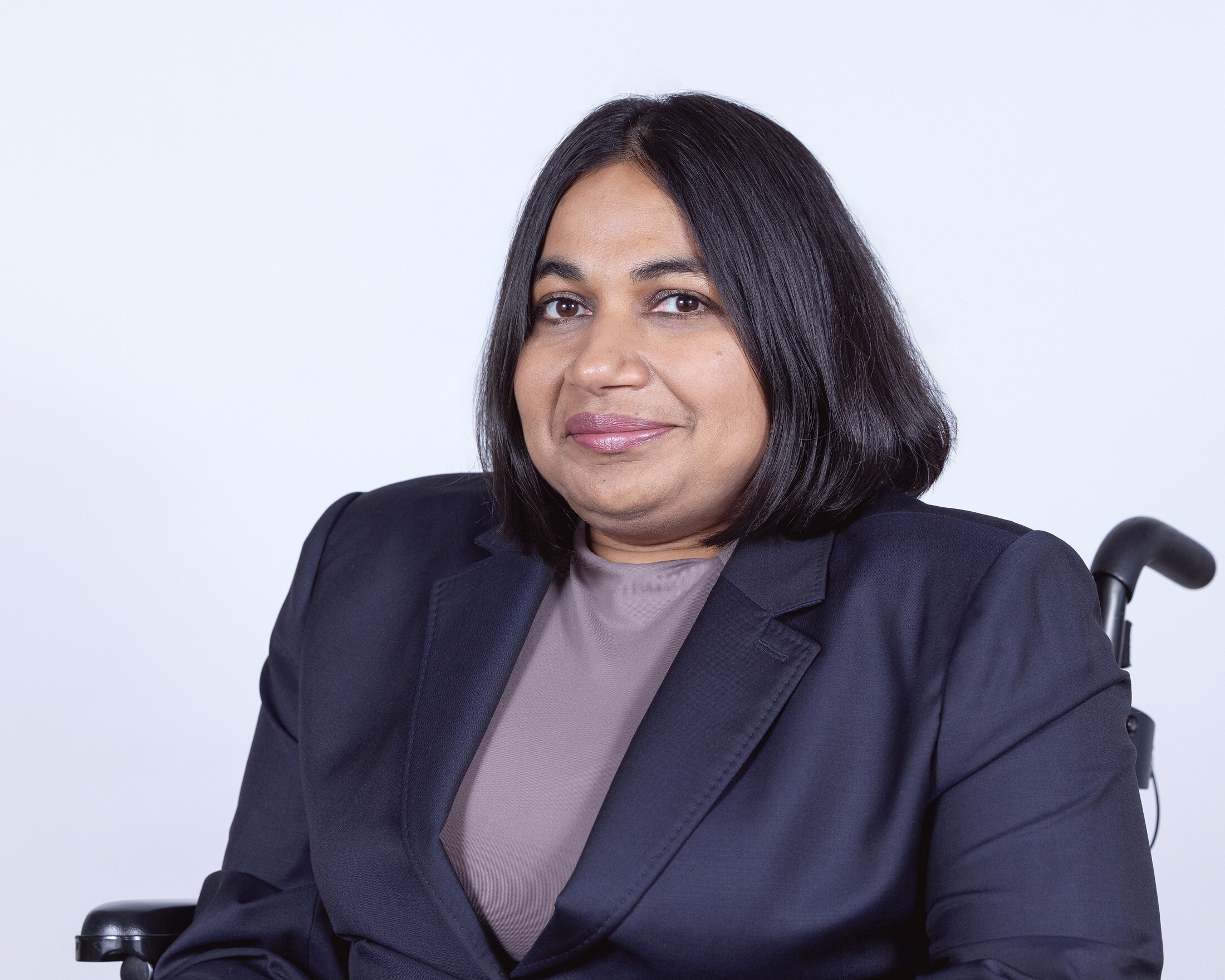 A person wearing a dark blazer and seated in a wheelchair against a light background.