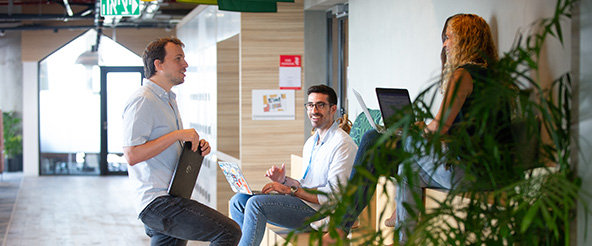 Three people having a casual meeting in a modern office space, sitting and standing near a hallway with laptops open, engaging in discussion.