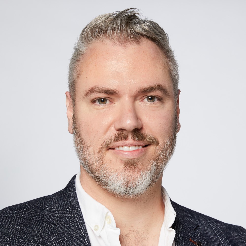 Professional headshot of a man with grey hair and beard wearing a blazer