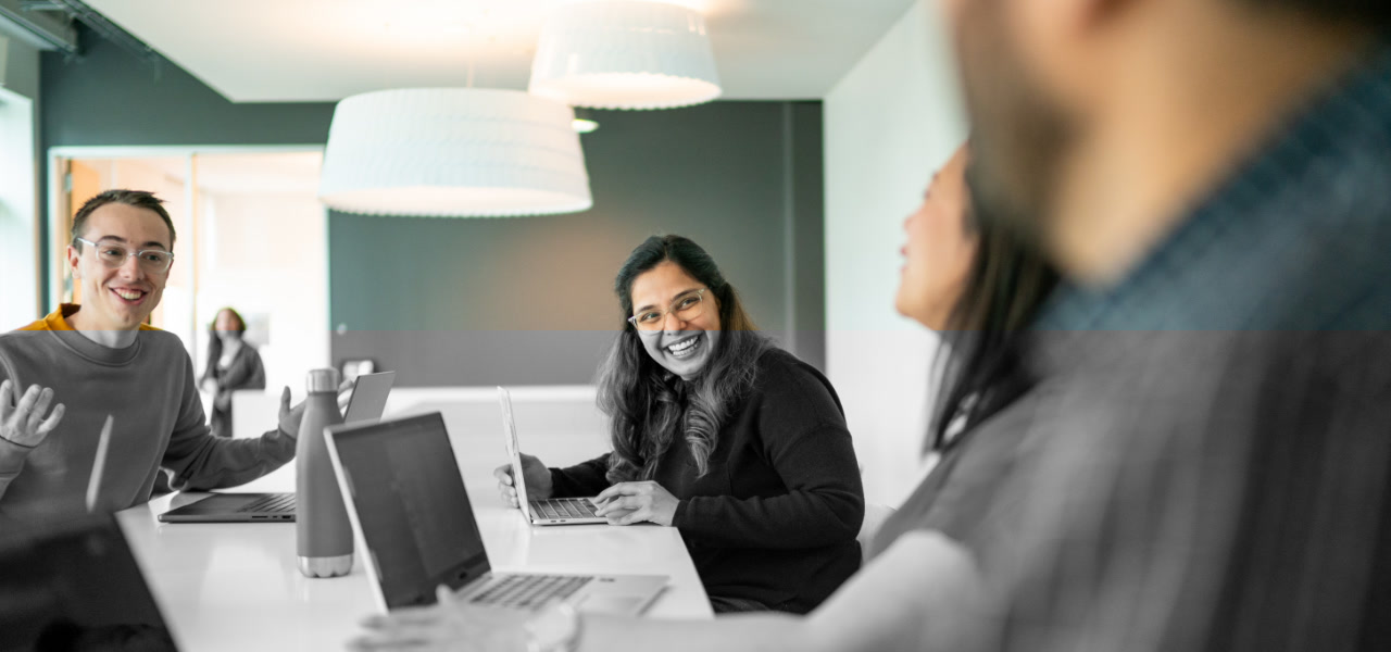 A group of AWS employees collaborating and smiling together around a table with laptops in a modern office environment.