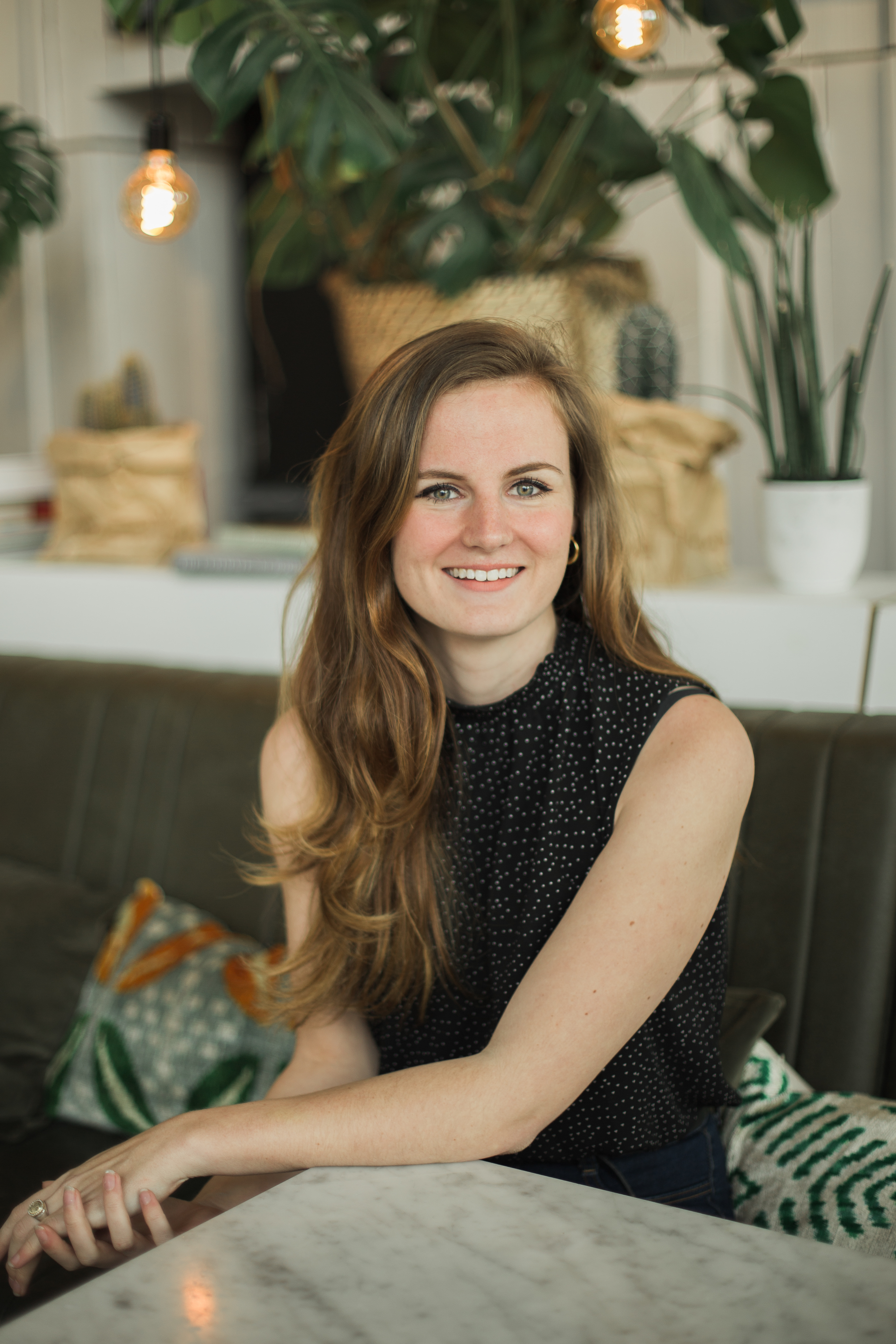 A woman with long hair, wearing a sleeveless black top, sits and smiles at a table in a cozy café. There are indoor plants and warm lighting in the background, creating a welcoming atmosphere.