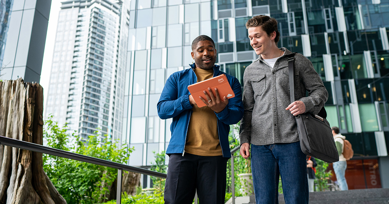 Two professionals standing outside an office building, looking at a laptop with an AWS logo. The modern cityscape, trees, and tall glass buildings are visible in the background.