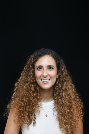 A woman with long curly hair smiling, photographed against a black background. She is wearing a sleeveless white top and a necklace.