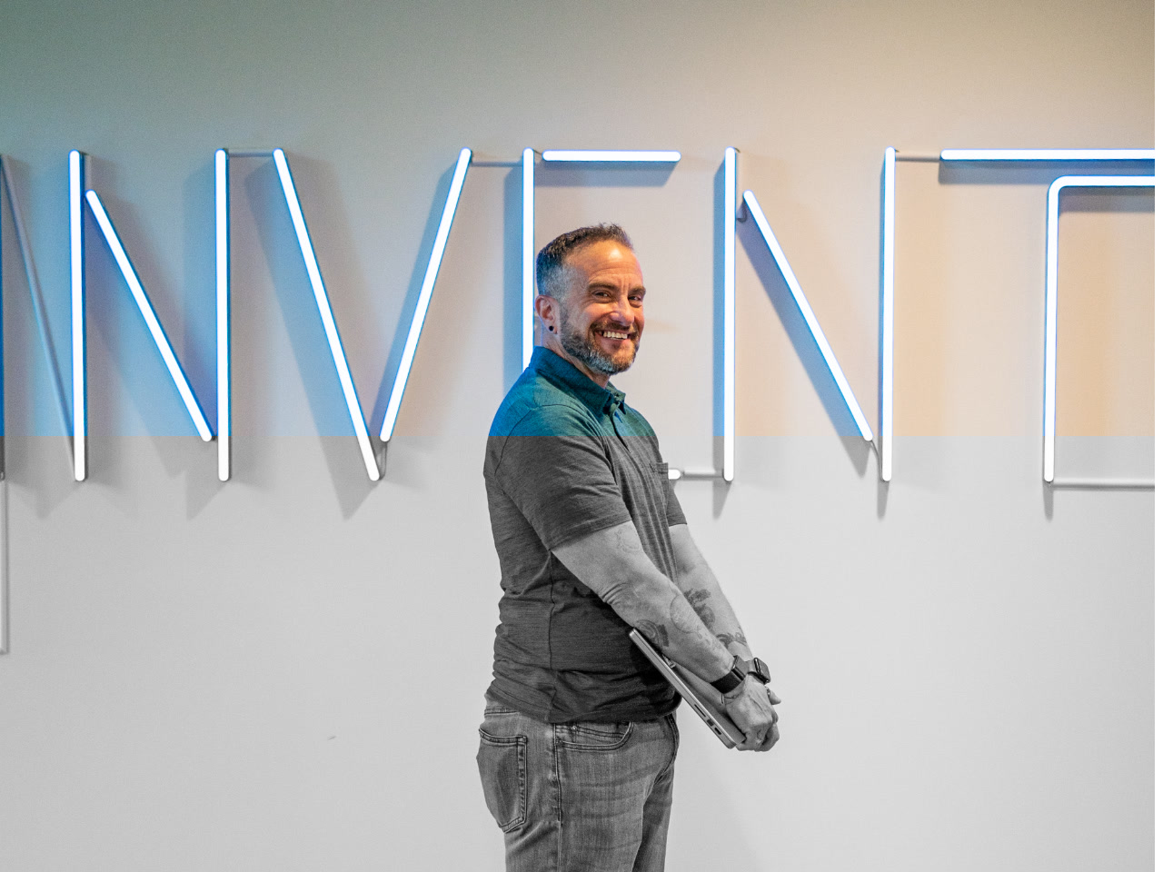 An AWS employee stands smiling and holding a laptop in front of a wall with illuminated 'INVENT' signage.