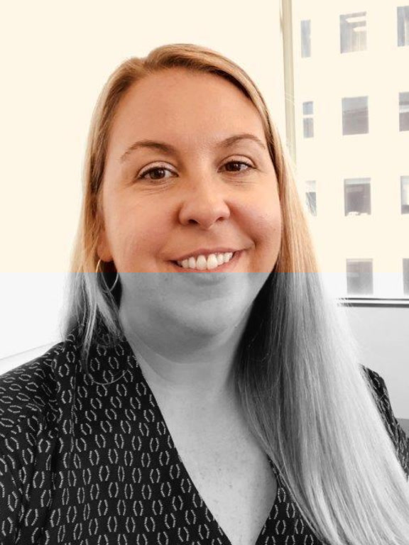 A person with long blonde hair wearing a patterned black top, sitting indoors near a window with a city building visible outside.