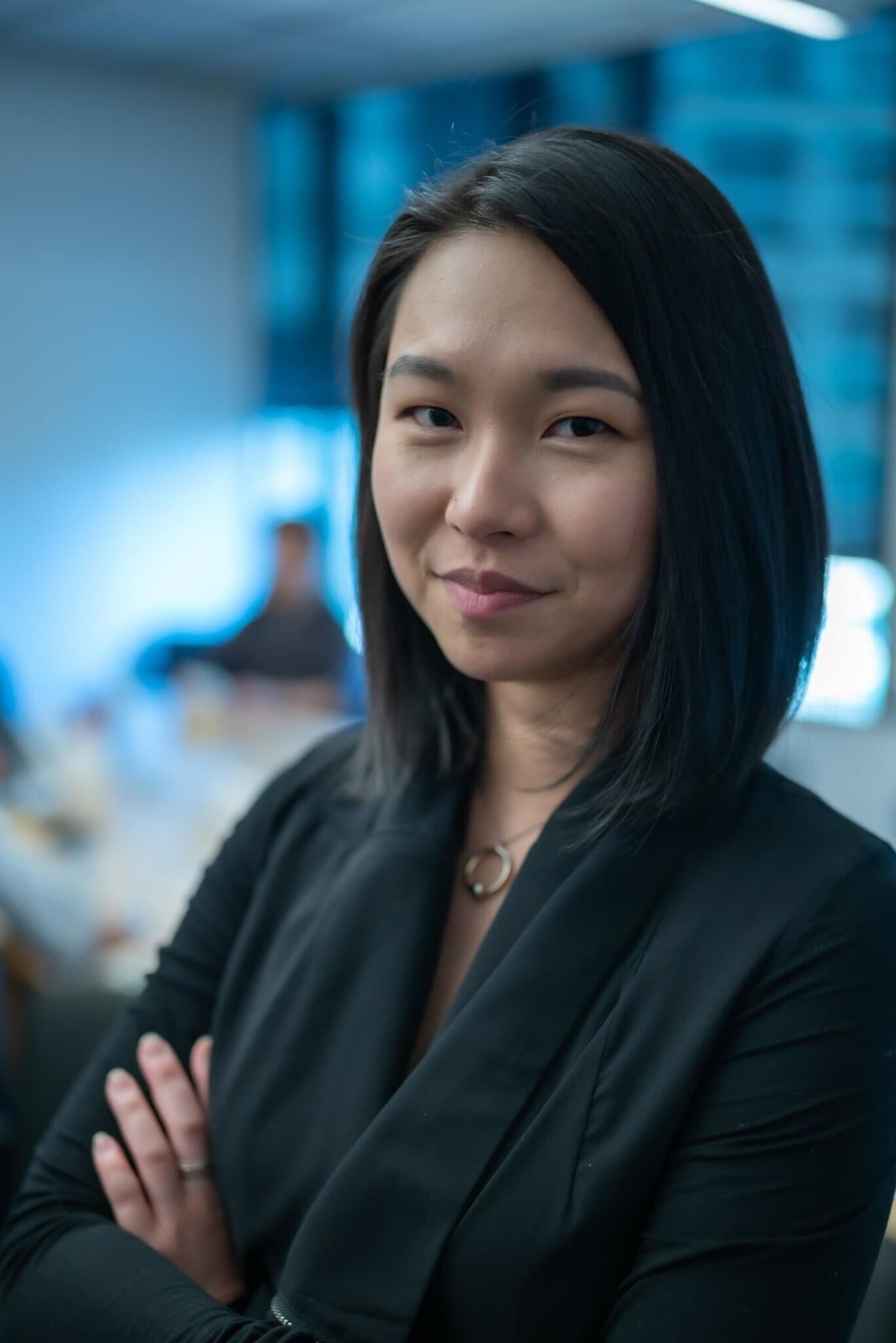 A professional woman with dark hair, wearing a black blazer, stands confidently in a modern office environment with blurred colleagues and windows in the background.