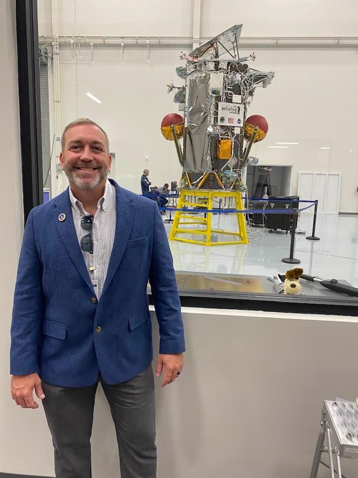 A man wearing a blue blazer is standing in front of a large window in a laboratory. Behind the window, a spacecraft with visible wiring and equipment is displayed on a yellow platform. The setting appears to be a clean, high-tech lab environment related to space or aerospace engineering.