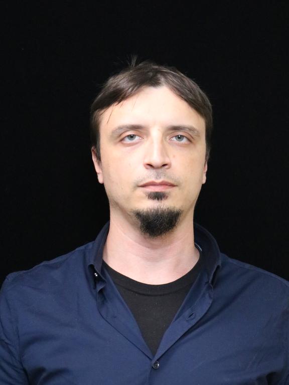A professional headshot of a person wearing a dark shirt, posed against a solid black background.