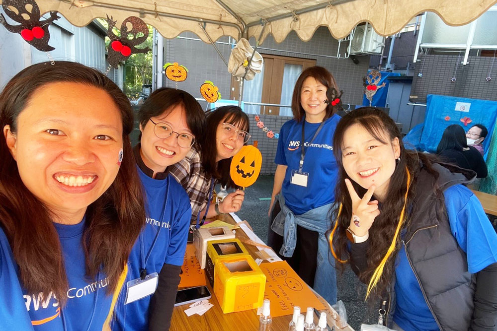 A group of smiling AWS Communities members posing together at a Halloween-themed event under a tent, wearing AWS Communities shirts and festive Halloween accessories.