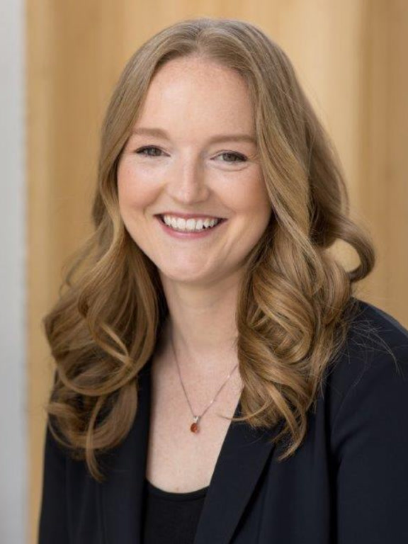 A person with long, wavy, light brown hair wearing a black blazer and a necklace with a small pendant.