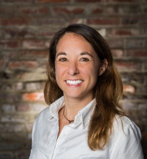 A person wearing a white shirt with long brown hair, standing in front of a textured brick wall.