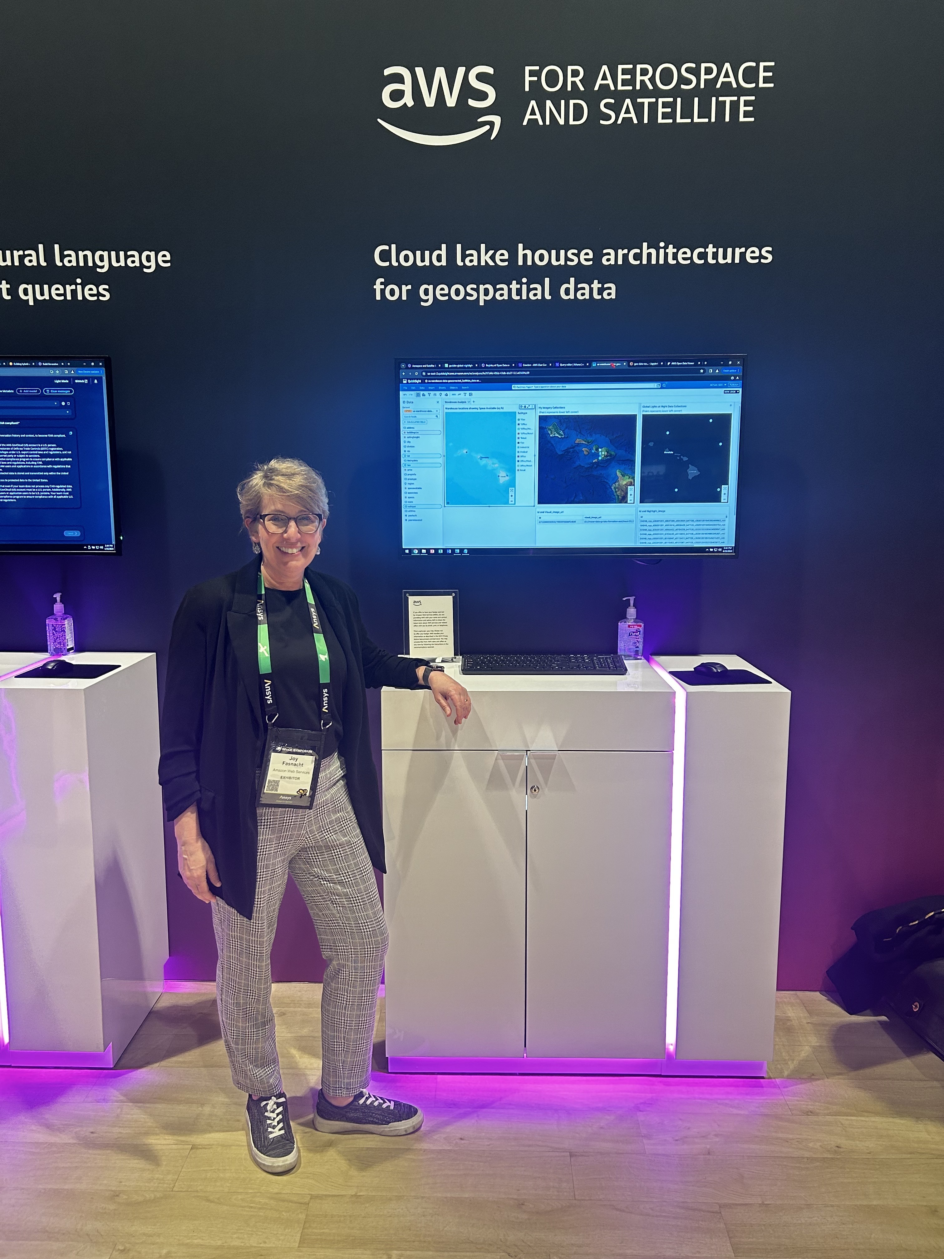 A person stands at an AWS Aerospace and Satellite booth featuring a cloud lake house architecture demo for geospatial data, with a large monitor displaying geospatial information.