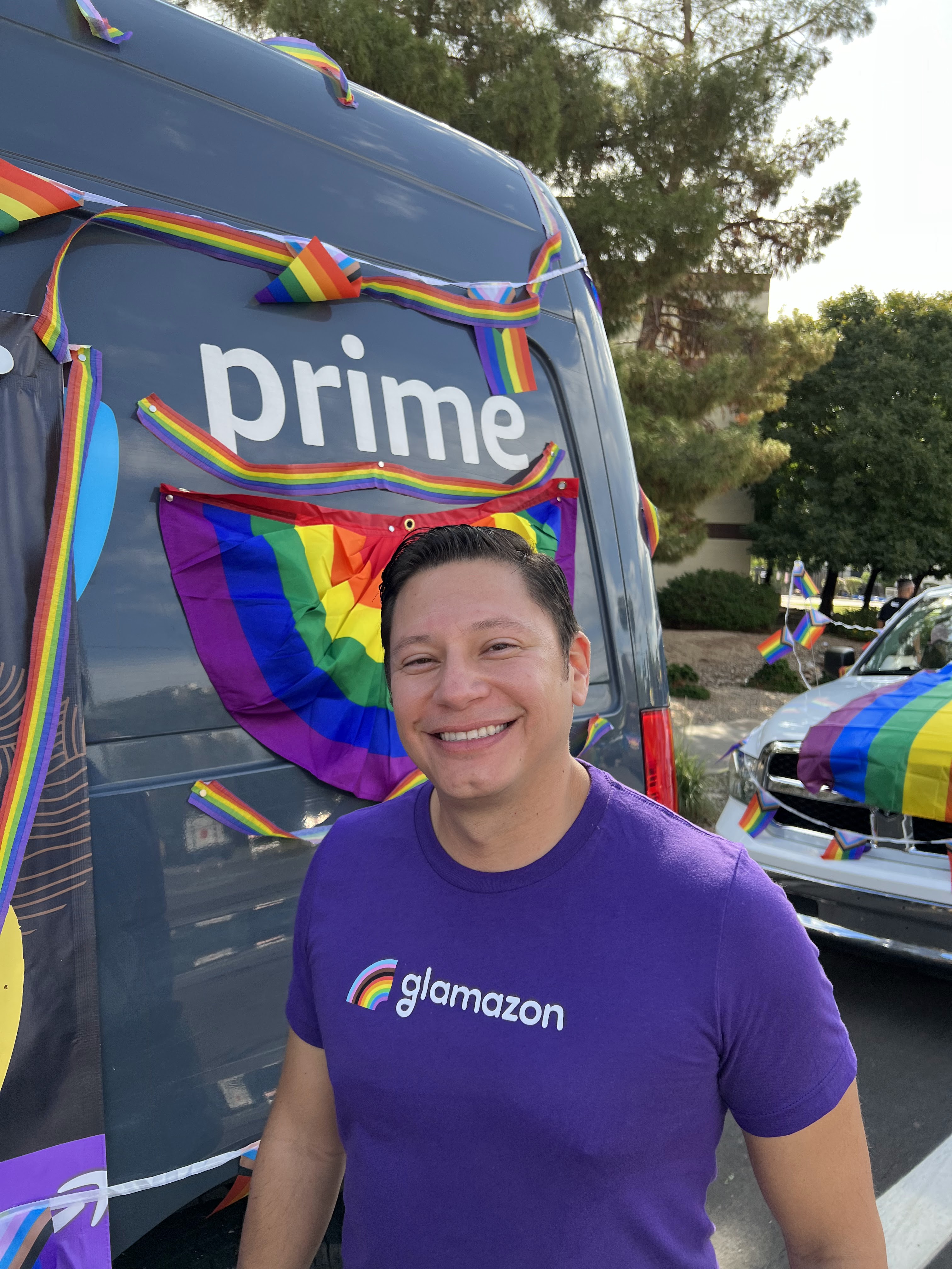 A smiling person wearing a purple 'glamazon' t-shirt, standing in front of an Amazon Prime delivery van decorated with rainbow pride flags and ribbons at a Pride parade.