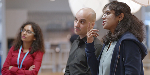 A group of AWS employees engaged in a discussion. The image features three people standing together in an office or collaborative workspace, with one person speaking while the others listen attentively.