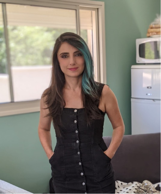 A woman with long dark hair featuring a blue streak, wearing a black dress, standing in a casual room with a light green wall, a couch, and a kitchen area in the background.