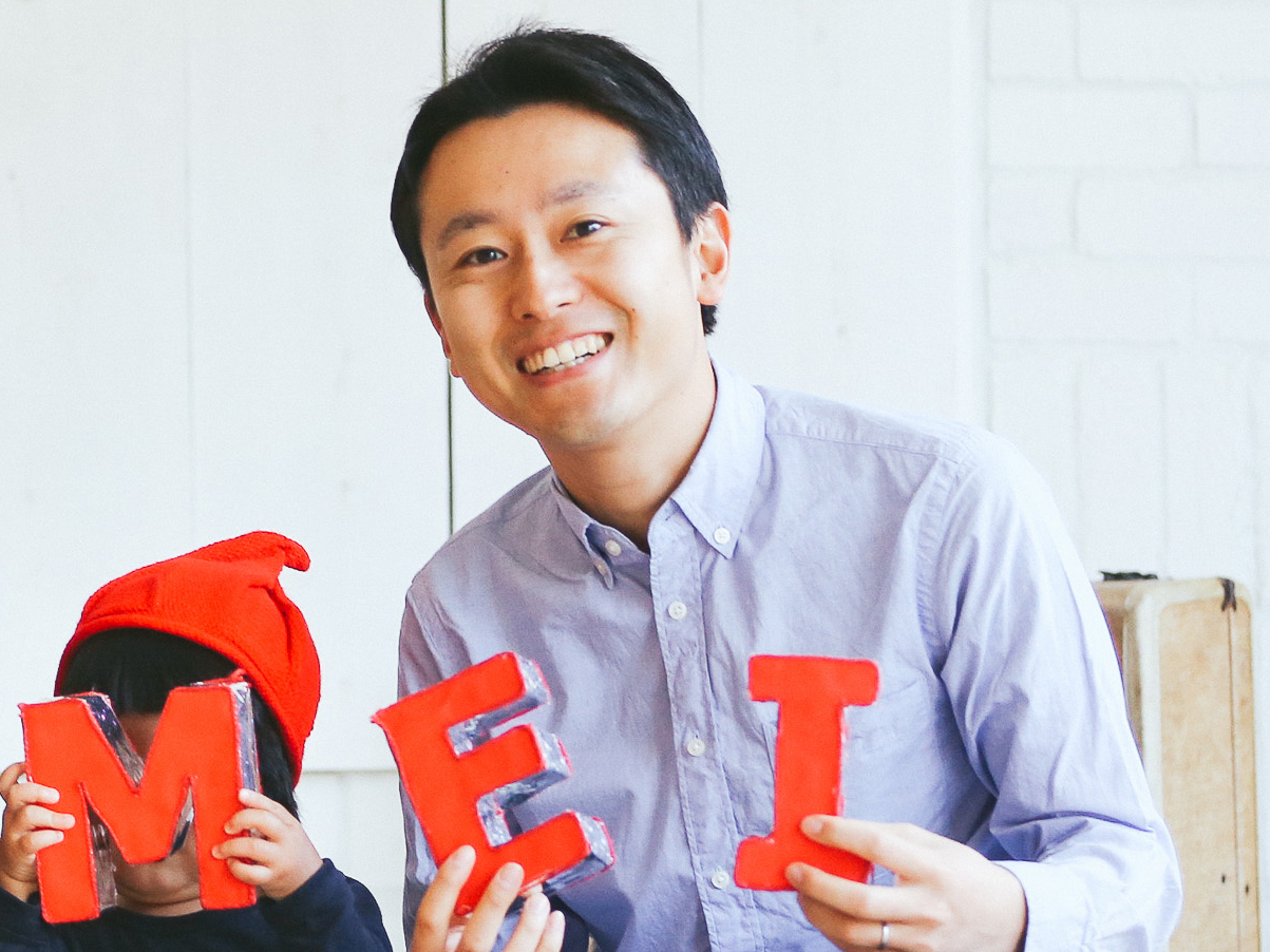A smiling man and a child, who is wearing a red hat, hold large red letters 'M', 'E', and 'I'. The child holds the 'M' and 'E' letters in front of their face, while the man holds the 'I' and smiles at the camera.