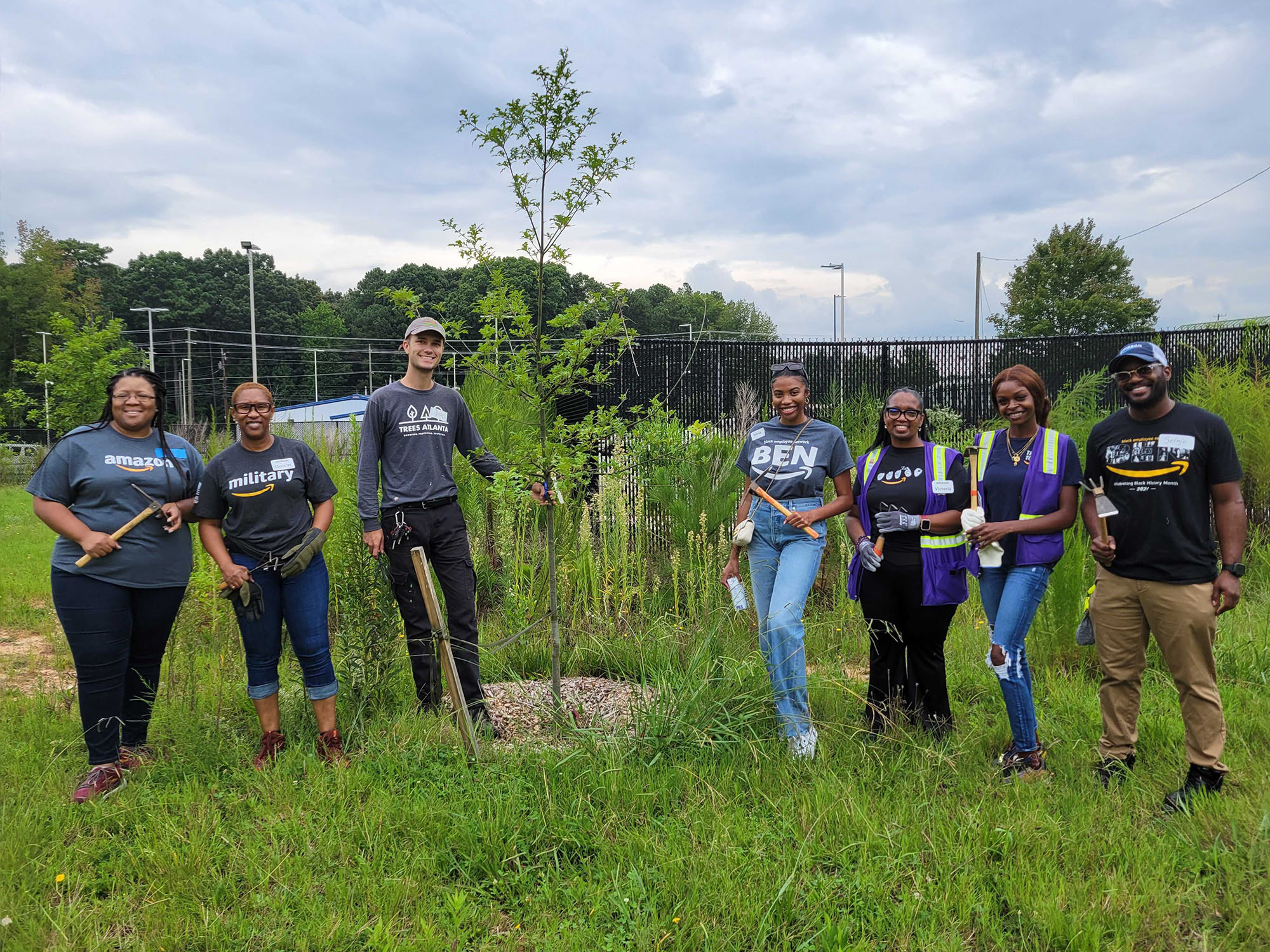 A group of Amazon employees volunteering outdoors, smiling and standing together while planting trees in a green garden area.