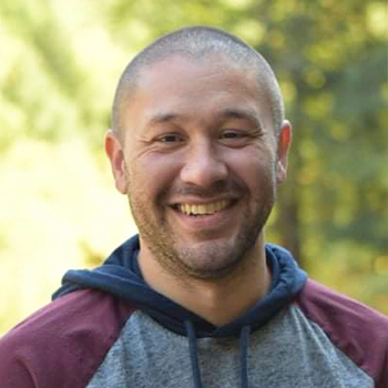 A close-up headshot of a smiling person wearing a hoodie, standing outdoors with a blurred background of greenery.