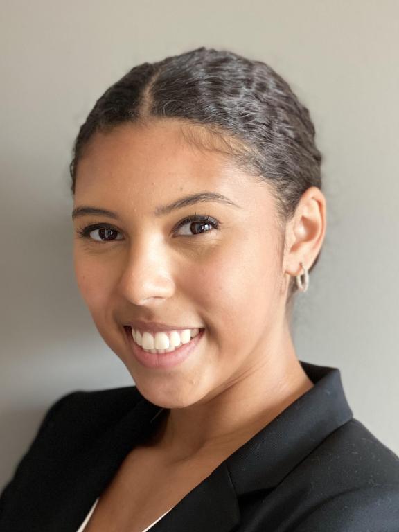 Professional headshot of a woman smiling, wearing a black blazer, with her hair neatly styled back, in front of a neutral background.