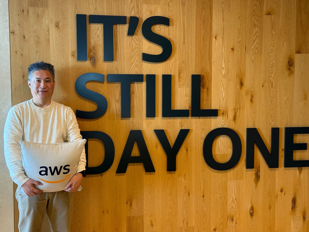 A person stands in front of a wooden wall with large black letters reading 'IT'S STILL DAY ONE,' holding a pillow with the AWS logo.