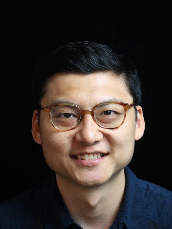 Portrait photo of a man wearing glasses, smiling, with a dark background.