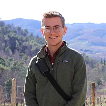 A person wearing glasses and a green jacket stands outdoors in front of scenic hills and trees, smiling at the camera.