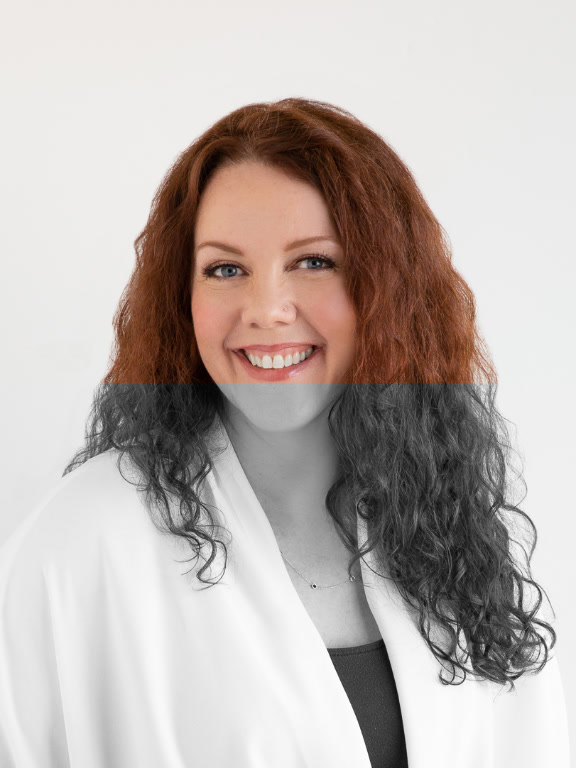 A person with long, curly red hair wearing a white top over a dark blue shirt against a plain background.
