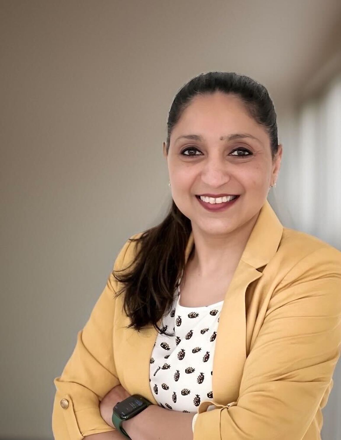 A professional portrait of a woman wearing a yellow blazer, smiling with arms crossed, and standing in a modern indoor setting.