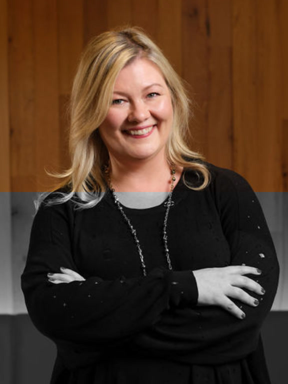 A person with long blonde hair wearing a black top and a necklace, standing with arms crossed against a wooden background.