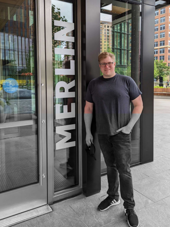 A person standing outside the entrance to the Merlin office building, with the building name visible on the glass door.