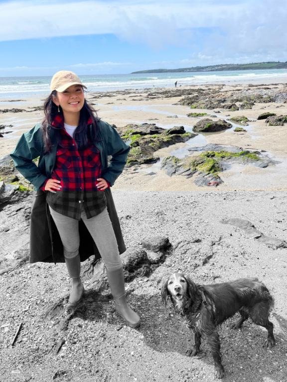 A person in a green raincoat and boots stands on a rocky beach next to a wet brown dog.