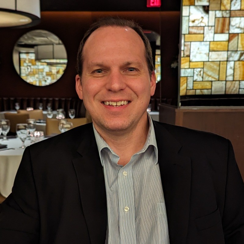 A person in formal attire sitting in a restaurant with round mirrors, stained glass, and set tables in the background.