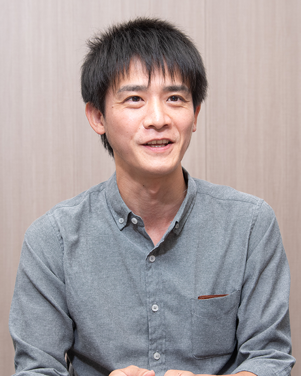 A man wearing a grey button-up shirt sits for an interview portrait against a plain background.