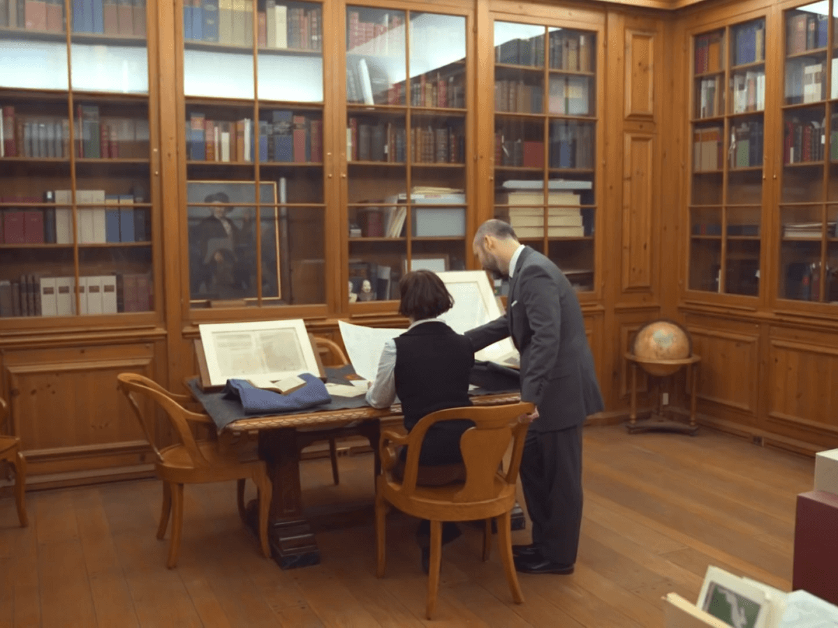 Two people working together at a computer in a library, surrounded by wooden bookshelves filled with books.