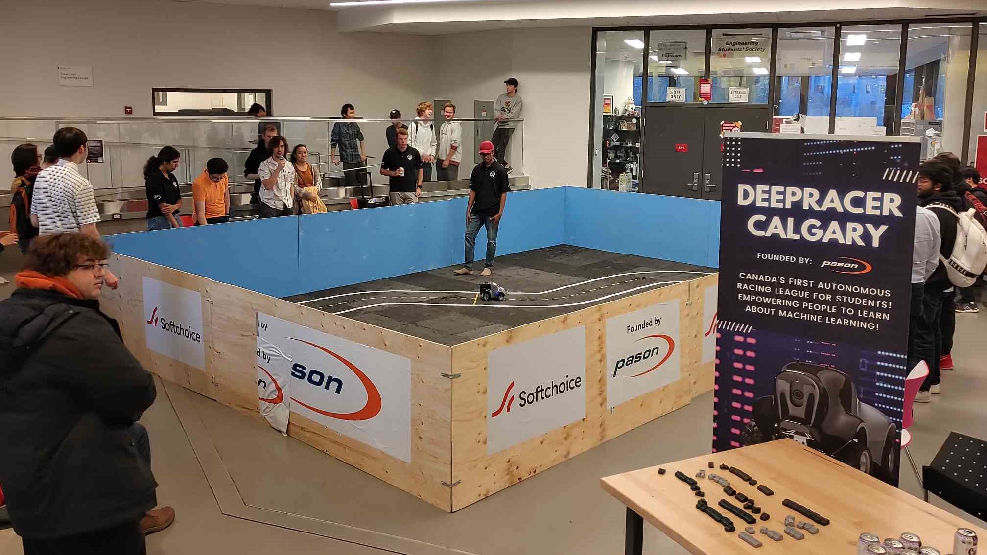 A group of students and spectators gather around an indoor autonomous racing track at the University of Calgary for a DeepRacer event. The event features AWS DeepRacer, an autonomous racing league designed to teach students about machine learning, with a model car navigating a track and signage promoting DeepRacer Calgary.