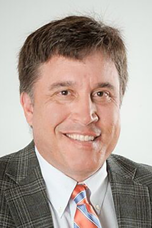 Portrait headshot of a male professional wearing a plaid suit jacket, white shirt, and striped tie, smiling at the camera with a neutral background.