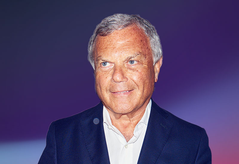 An older man with gray hair wearing a dark suit jacket and white shirt, set against a purple gradient background.