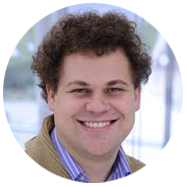 2A person with curly brown hair wearing a striped shirt and a tan jacket, set against a blurred background.