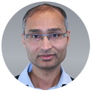 2A person wearing a light blue collared shirt and a black sweater against a gray background.