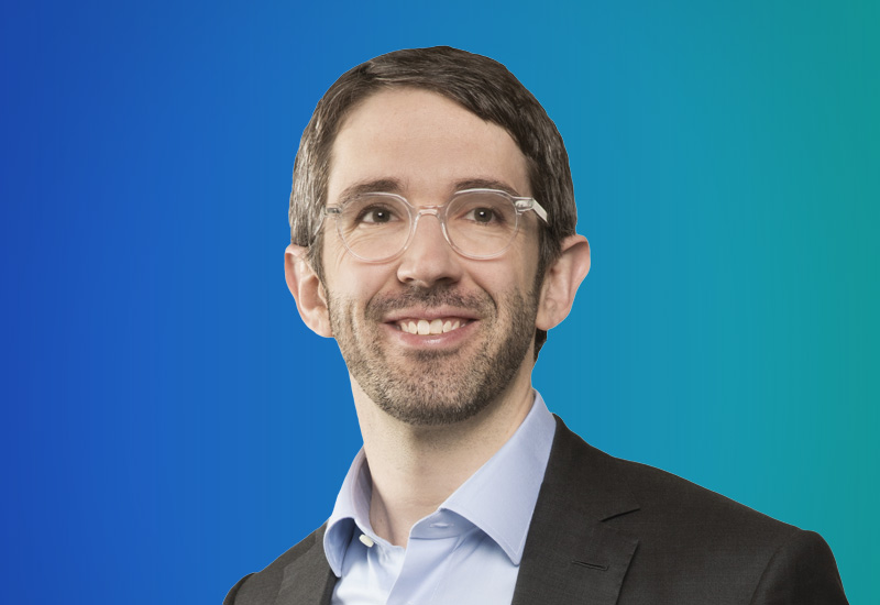 A person wearing a suit and light blue shirt against a gradient blue background.