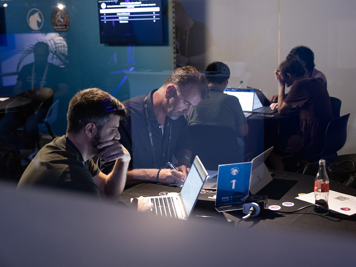 People working collaboratively on laptops at a table during an event, with a sign labeled "1" and a Coca-Cola bottle visible.