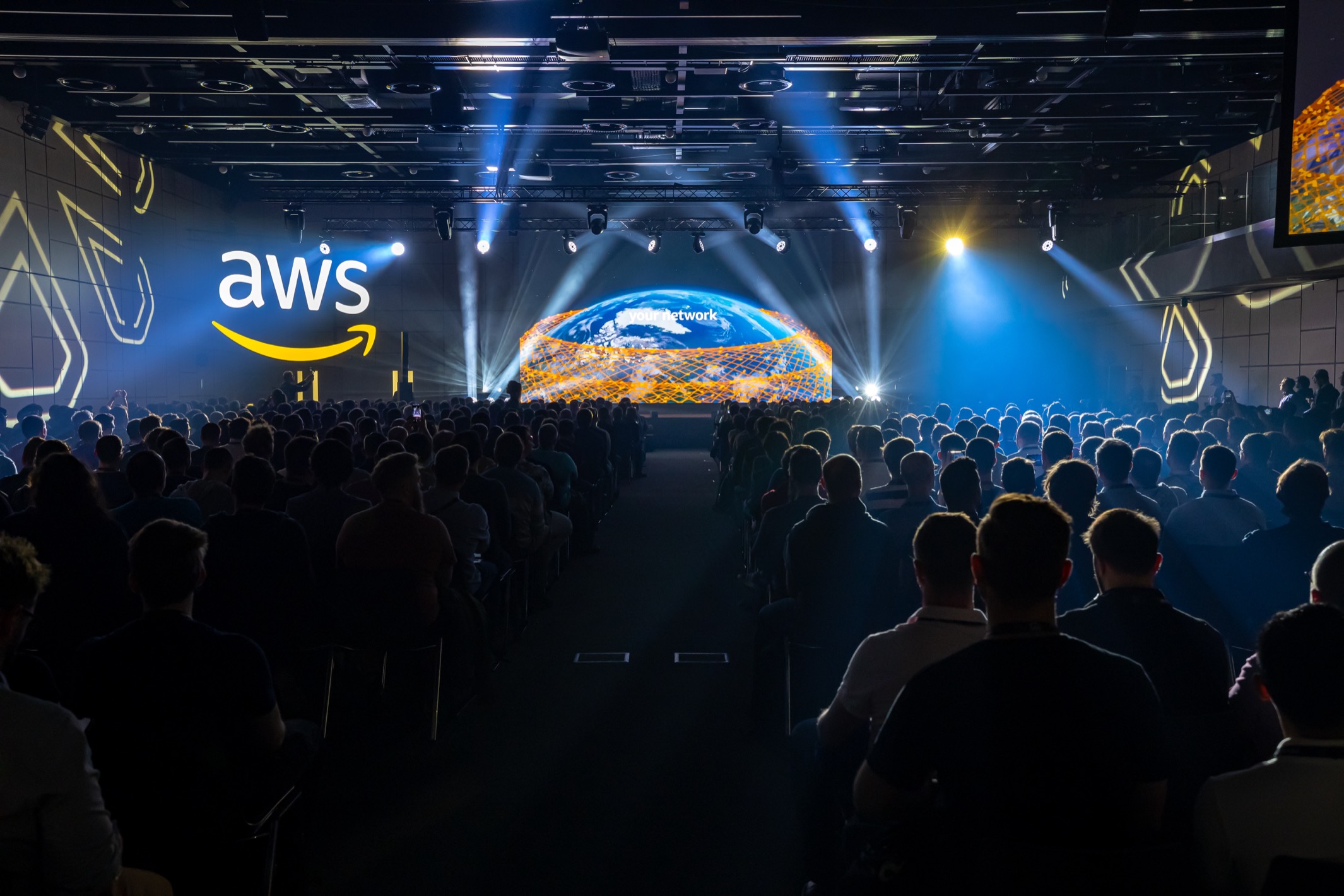 A large audience attends an AWS Cloud Days 2024 conference in Prague, viewed from the back of the room looking toward the stage with bright lights, the AWS logo, and an illuminated globe on the main screen.