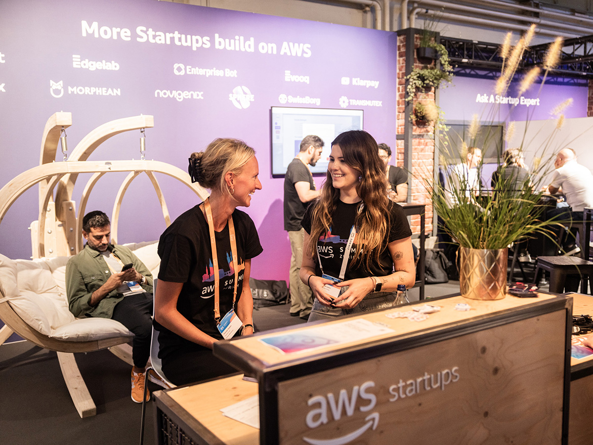 People interacting at an AWS Startups booth with a modern lounge chair and a purple backdrop featuring startup logos.