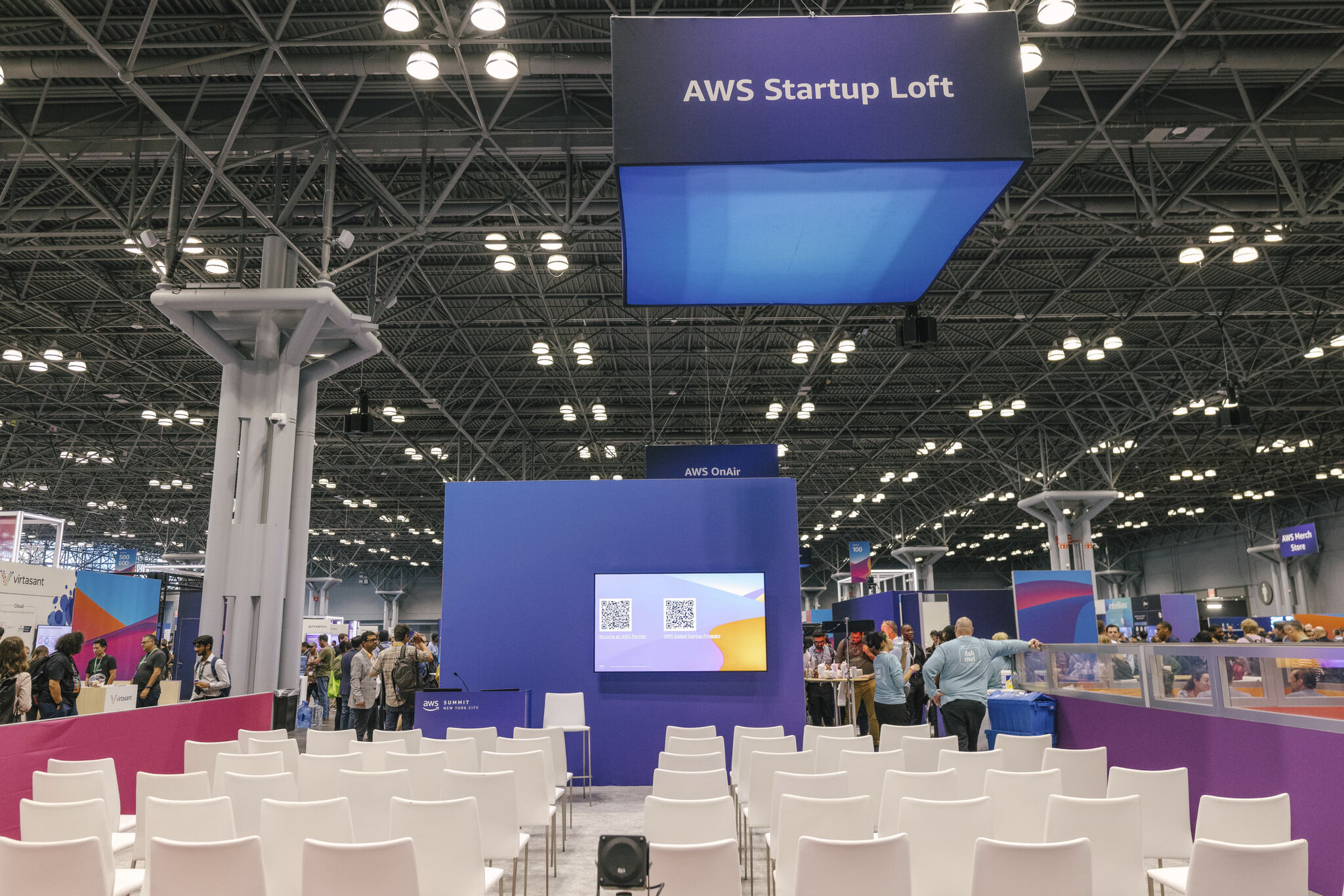 A conference setup at the AWS Startup Loft with rows of white chairs facing a screen displaying QR codes, surrounded by exhibition booths and attendees.